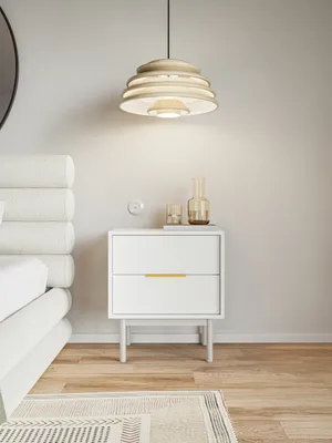 A white nightstand with two drawers and a gold handle is placed next to a bed in a bedroom. A pendant light hangs above the nightstand.