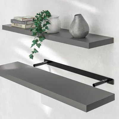 Two gray shelves mounted on a white wall. The top shelf has books, a plant, and a vase. The bottom shelf has a black bracket.