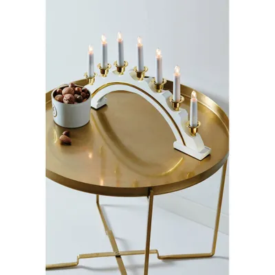 A white and gold candelabra with seven lit candles sits on a round, gold-colored table. A small white bowl filled with nuts is next to the candelabra, with a few nuts scattered on the table.