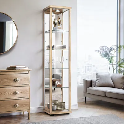 A tall glass display cabinet with wooden trim, filled with decorative items, stands in a living room next to a chest of drawers and a sofa.