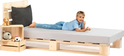 A young boy is lying on a bed made of wooden pallets, with a mattress and a pillow. There are also some stuffed animals on a wooden crate next to the bed.