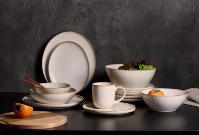 A collection of ceramic tableware, including plates, bowls, and a mug, arranged on a dark surface against a dark gray textured background. A persimmon is placed on a wooden cutting board in the foreground.