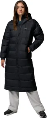 A woman wearing a long black puffer coat and light grey sweatpants stands against a white background.