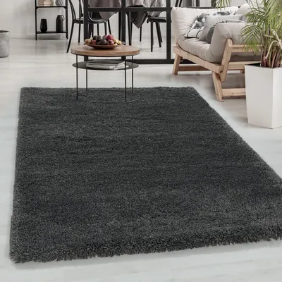 A dark gray shaggy rug is shown in a living room setting with a coffee table, chairs, and a sofa in the background.