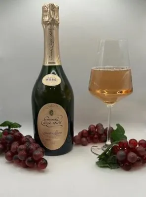 A bottle of rosé sparkling wine and a glass of rosé wine, with grapes on the side, on a plain background.