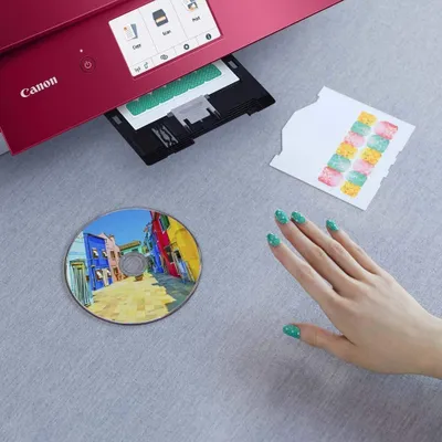 A red Canon Pixma printer is shown printing a sheet of paper, with a CD and a hand with decorated nails nearby.