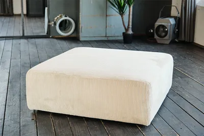 A cream-colored corduroy ottoman sits on a dark wooden floor in a studio-like setting with industrial elements in the background.