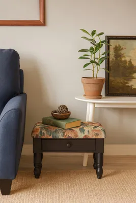 A close-up shot of a decorative ottoman placed next to a blue sofa and a side table with a potted plant and a framed painting.