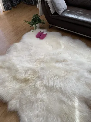 A fluffy white sheepskin rug is laid out on a wooden floor in front of a dark leather sofa. Two pink slippers are placed on the rug, and a small potted plant is visible in the background.
