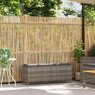 A garden bench with cushions is placed on a patio in front of a bamboo fence, with plants and trees in the background.