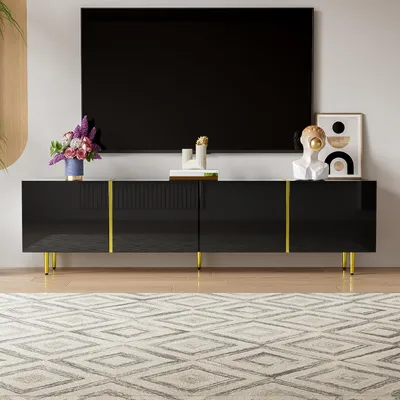 A modern black TV stand with gold accents is shown in a living room setting, with a large black TV mounted on the wall above it. A rug with a geometric pattern is in the foreground.