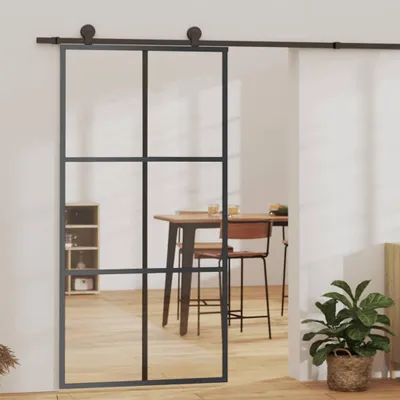 A sliding glass door with a black frame is shown in a room with wooden floors. A dining table and chairs are visible through the glass, and a potted plant is in the foreground.