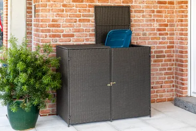 A dark gray woven storage bin with a blue shovel inside, placed on a patio next to a potted plant.