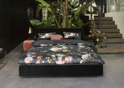 A bedroom scene with a bed featuring floral bedding, plants, and stairs in the background.