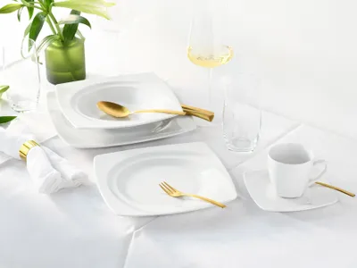 A white porcelain dinnerware set is arranged on a white tablecloth, with gold cutlery and a green vase with flowers in the background.