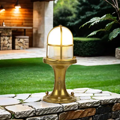 A brass outdoor lamp is placed on a stone wall in a garden setting, with a wooden patio and greenery in the background.