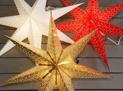 Three illuminated paper star lanterns in white, red, and gold are displayed against a gray wooden background.