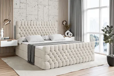 A bedroom scene featuring a large, tufted boxspring bed with a headboard, mattress, and topper. The bed is dressed with white pillows and a gray throw blanket. Two modern white nightstands with lamps are placed on either side of the bed. The background inc
