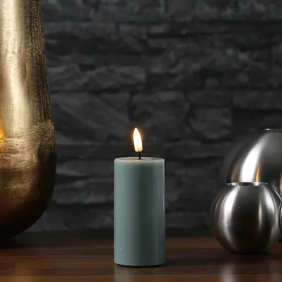 A lit candle sits on a wooden surface in front of a dark stone wall, with a large gold vase on the left and silver decorative spheres on the right.