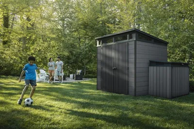 A family is enjoying a sunny day in their backyard, with a young boy playing soccer near a modern shed.