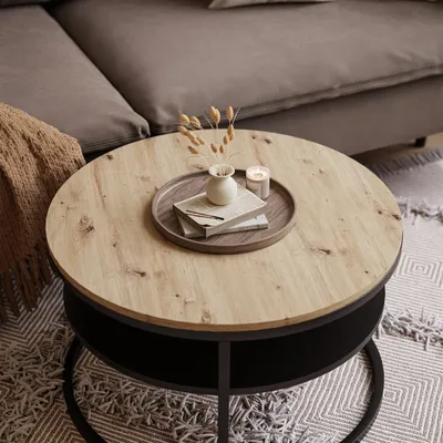 A round coffee table with a wooden top and a dark base, placed on a patterned rug next to a sofa. Decorative items include a small vase with dried flowers, a candle, and books on a tray.