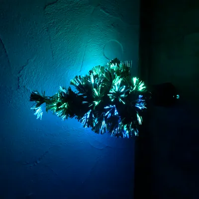 A small, illuminated artificial Christmas tree with fiber optic lights in green and blue, set against a textured wall.