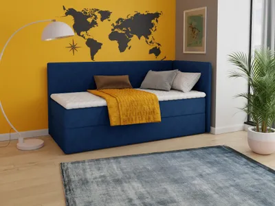 A bedroom scene with a dark blue boxspring bed, a yellow blanket, and pillows. A world map is on the yellow wall behind the bed. A floor lamp is on the left, and a potted plant is on the right.