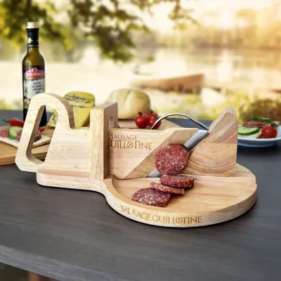 A wooden sausage guillotine is set up outdoors on a table, with food items like cheese, bread, and tomatoes in the background, suggesting a picnic or outdoor meal setting.