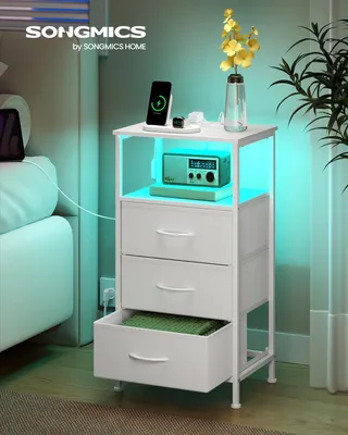 A white nightstand with drawers and an open shelf, featuring a phone charging, a radio, and a vase with flowers. The nightstand has integrated LED lighting.