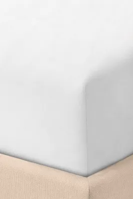 A close-up view of a white fitted sheet on a bed, with a beige bed skirt visible at the bottom.