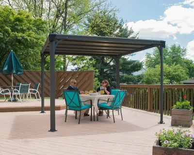A couple is enjoying a meal under a metal pergola on a wooden deck, surrounded by trees and a wooden fence.