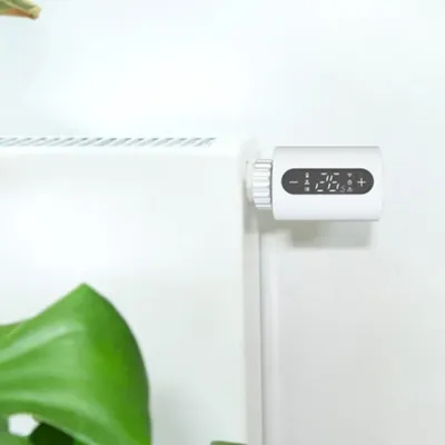 A white smart thermostat head attached to a white radiator, with a large green leaf in the foreground.