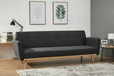 A dark gray sofa with wooden legs is shown in a well-lit room with two framed pictures on the wall and a small side table with a lamp.