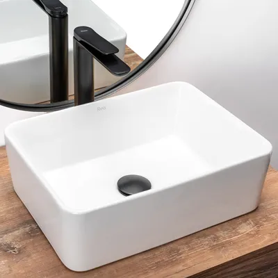 A white rectangular basin sink with a black drain and a black faucet, placed on a wooden surface. A round mirror reflects the faucet and sink.