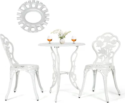 A white metal bistro set with two chairs and a table, decorated with rose patterns. The table has two glasses of amber liquid and a small vase of white flowers.