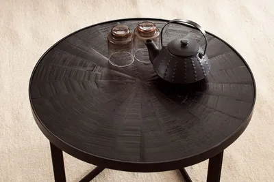 A round black coffee table with a textured surface, two glasses, and a black teapot on top, placed on a light beige carpet.