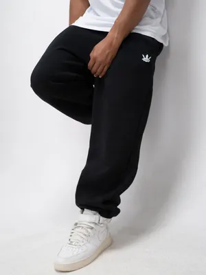 A person wearing black sweatpants and white sneakers, leaning against a white wall.