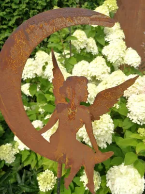 A rusty metal garden stake shaped like a fairy sitting on a crescent moon, set against a backdrop of white hydrangeas.