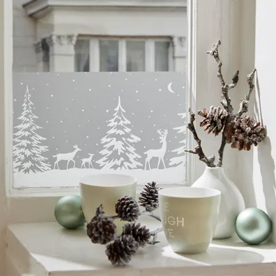 A festive window decoration with a winter forest and reindeer motif, set on a windowsill with decorative elements.