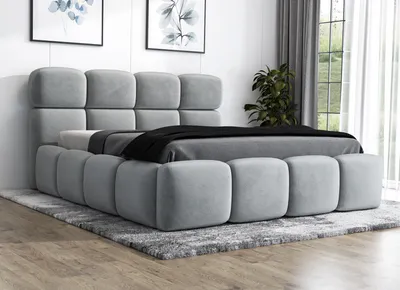 A modern, upholstered bed with a tufted headboard and frame, set on a rug in a room with artwork and curtains.