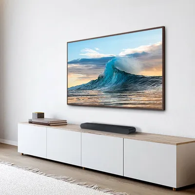 A soundbar is placed on a TV stand in front of a wall-mounted television displaying a wave.
