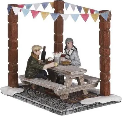 Two miniature figures are seated at a picnic table under a canopy decorated with bunting, with snow on the ground.