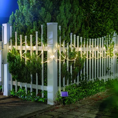 A white picket fence decorated with a string of warm white LED lights at night, with green bushes and a solar panel visible.