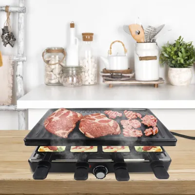 A raclette grill with meat and cheese on a wooden table, with kitchen items in the background.