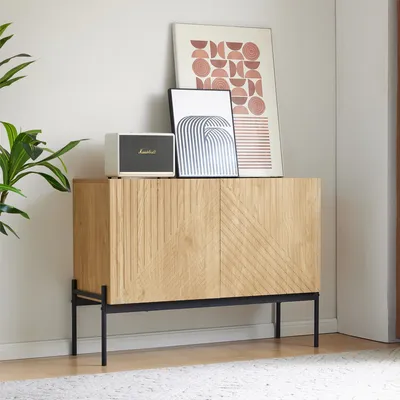 A wooden sideboard with black metal legs, decorated with two framed art prints and a speaker, placed on a rug in a room.