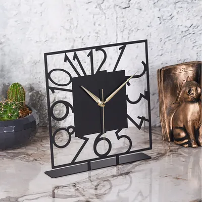 A decorative square clock with a modern design is placed on a marble surface, next to a potted cactus and a bronze cat statue.