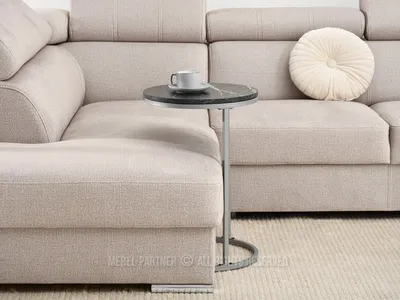 A small round side table with a marble top and metal base is placed next to a large, light-colored sectional sofa. A white coffee cup and saucer sit on the table. A round, tufted cushion is on the sofa.