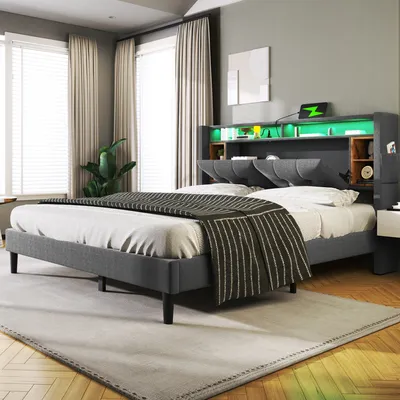 A modern bedroom with a grey upholstered bed frame, featuring a headboard with built-in storage, LED lighting, and a USB charging port. The bed is made up with a striped throw blanket and white bedding. The room has large windows with beige curtains and a 