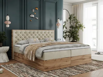 A bedroom scene featuring a large boxspring bed with a tufted headboard and wooden base, decorated with pillows and a throw blanket. The room has dark teal walls, decorative wall art, and a large plant near a window.