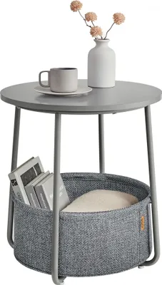 A round side table with a storage basket underneath, holding books and a cushion. A mug and a vase with flowers are on top.